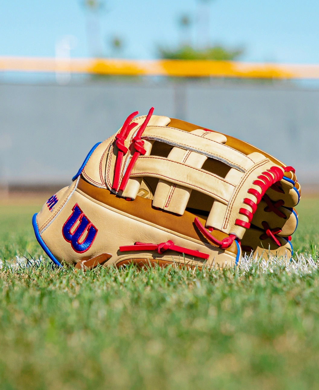 A Wilson A2000 1777 12.75″ Baseball Glove (WBG06001RT1275) in tan and brown Pro Stock leather, accented with bold red laces, lies on the grass near the infield, with a blurred fence and blue sky serving as the backdrop.