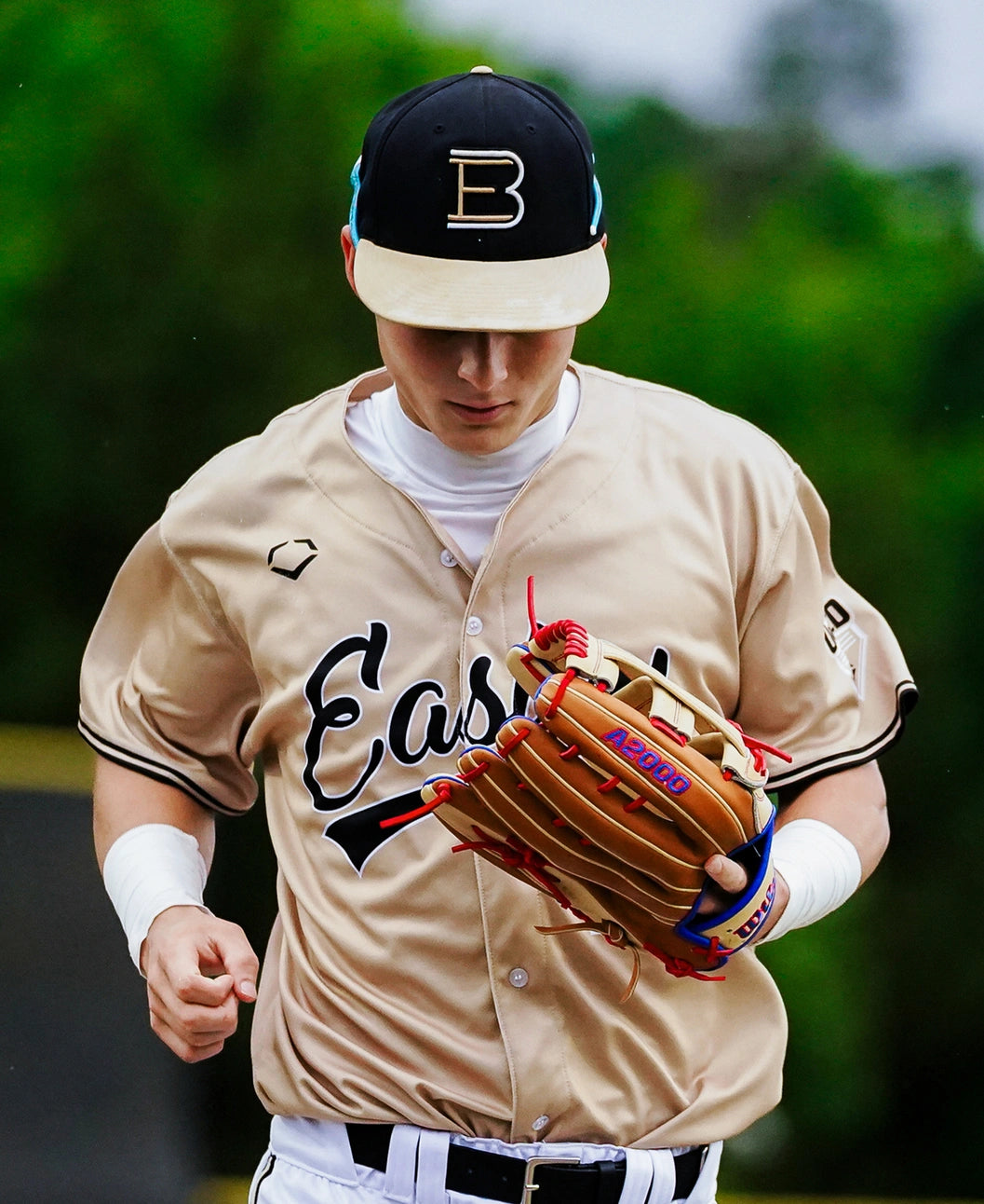 A baseball player in a tan Eason jersey and black cap with a gold B holds a Wilson A2000 1777 12.75″ Baseball Glove (WBG06001RT1275), crafted from Pro Stock leather, while walking on a field with a green blurred background.