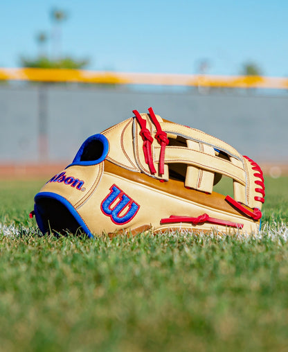A Wilson A2000 1777 12.75″ baseball glove (WBG06001RT1275) in tan Pro Stock leather, featuring red and blue accents, rests on vibrant green grass with a blurred field and fence behind it beneath a clear blue sky.