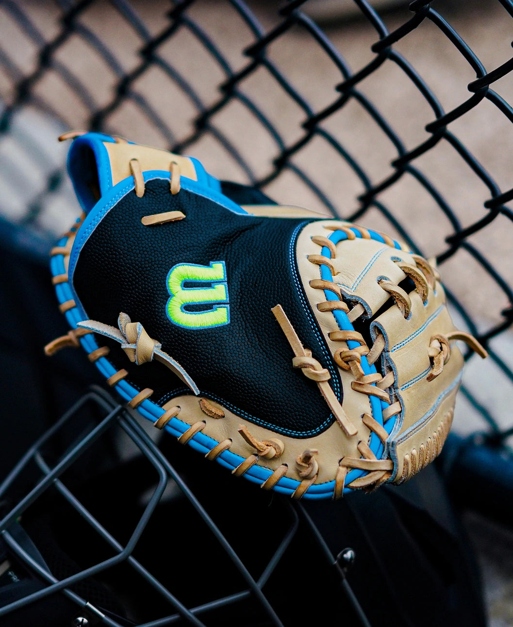 A Wilson A2000 M23SS 33.5″ Baseball Catcher’s Mitt (WBG06201RT335), featuring a tan and black ultra-light design with blue accents and a neon green W logo, rests on a metal fence near a baseball field.