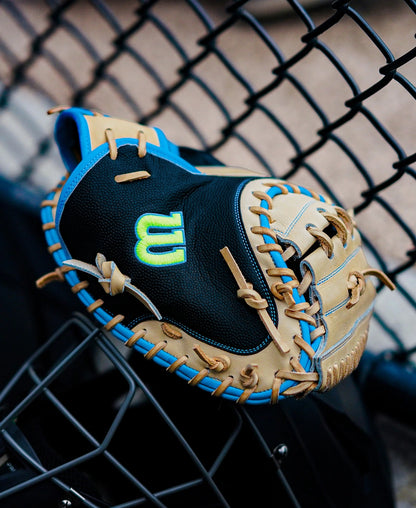 A Wilson A2000 M23SS 33.5″ Baseball Catcher’s Mitt (WBG06201RT335), featuring a tan and black ultra-light design with blue accents and a neon green W logo, rests on a metal fence near a baseball field.