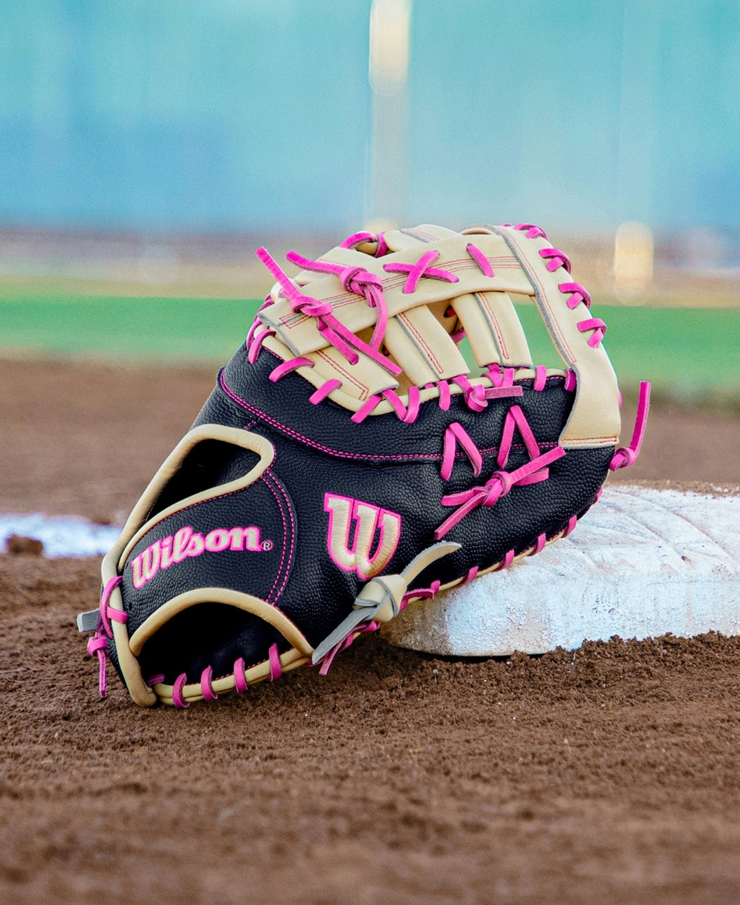 A Wilson A2000 1673SS 13″ Baseball First Base Mitt (WBG06301RT13) by Wilson, featuring a black and tan design with pink laces, rests on a baseball base on a dirt field, while green grass and a blue sky appear blurred in the background.