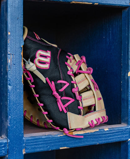 A Wilson A2000 1673SS 13″ Baseball First Base Mitt (WBG06301RT13) in black, tan, and pink rests on a blue wooden shelf. The glove features pink laces and a prominent Wilson “W” logo on the side.