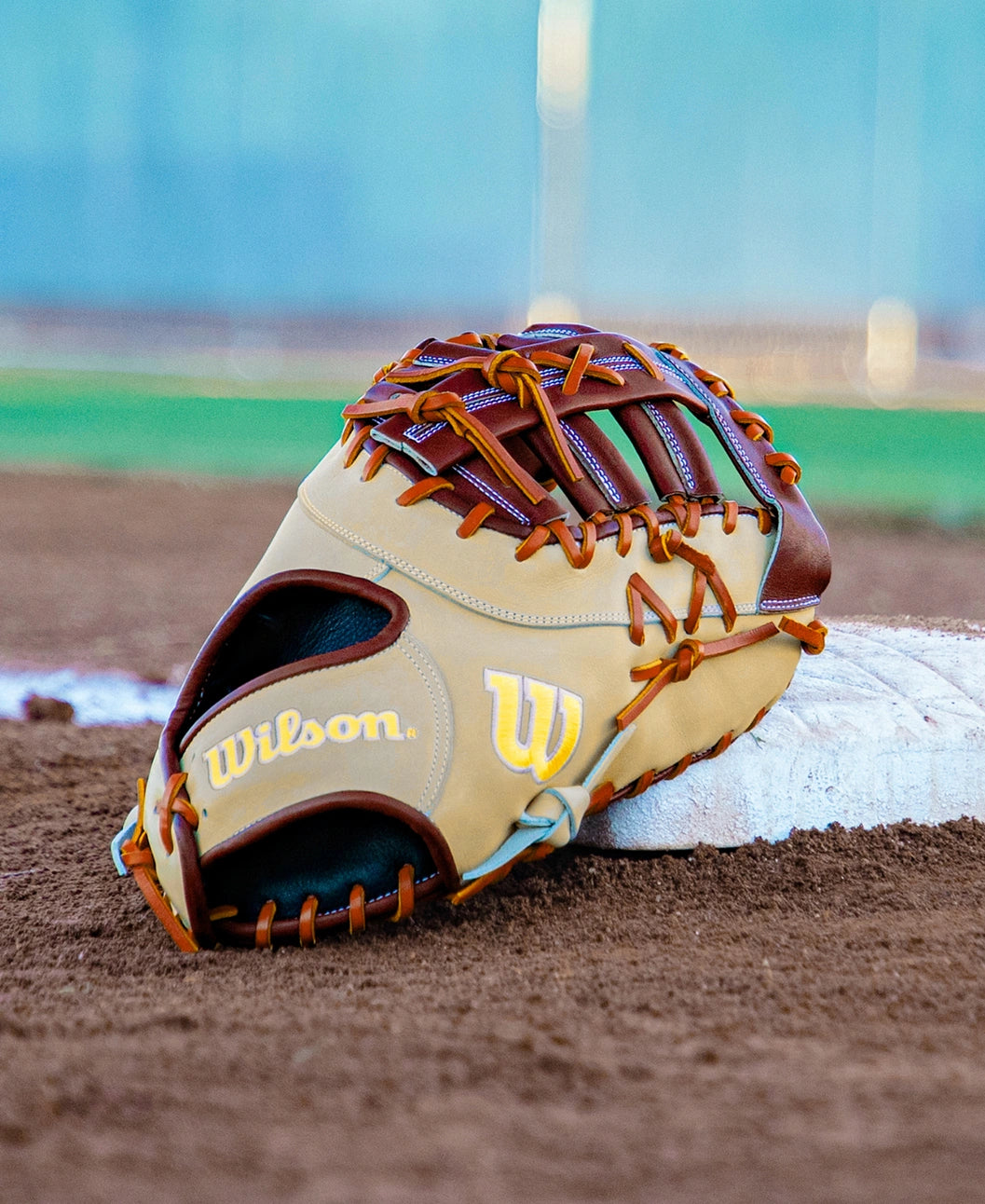 A Wilson first base mitt crafted from Pro Stock leather in tan and brown rests on the dirt by a white base on a baseball field, with green grass and a blurred fence in the background.