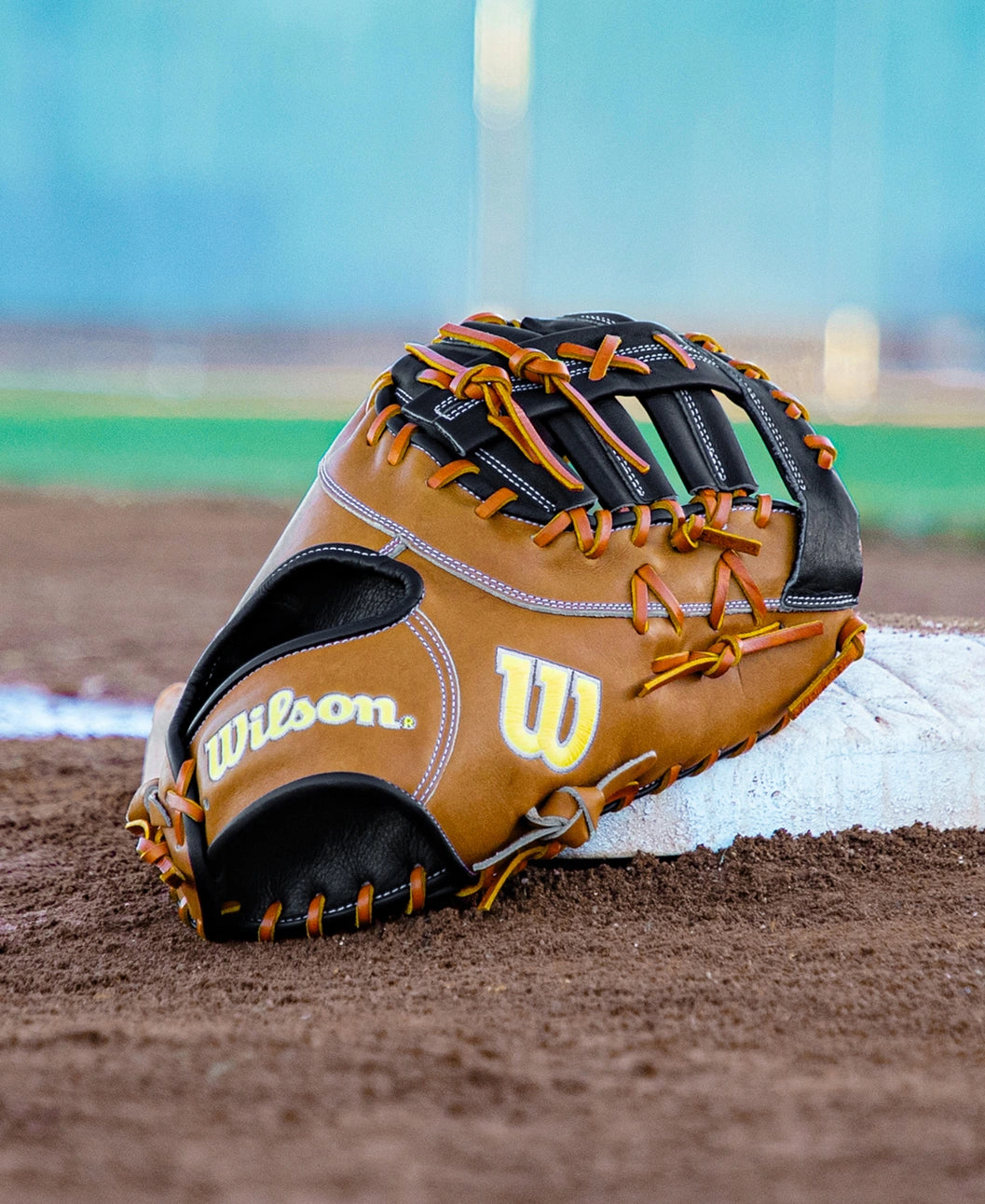 A Wilson A2000 1677 12.5″ Baseball First Base Mitt (WBG06501RT125) from Wilson, featuring durable Pro Stock leather in brown, lies on the dirt near a base on a baseball field, with green grass and a blue background in view.