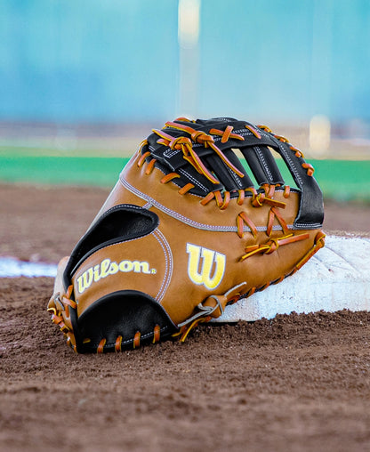 A Wilson A2000 1677 12.5″ Baseball First Base Mitt (WBG06501RT125) from Wilson, featuring durable Pro Stock leather in brown, lies on the dirt near a base on a baseball field, with green grass and a blue background in view.