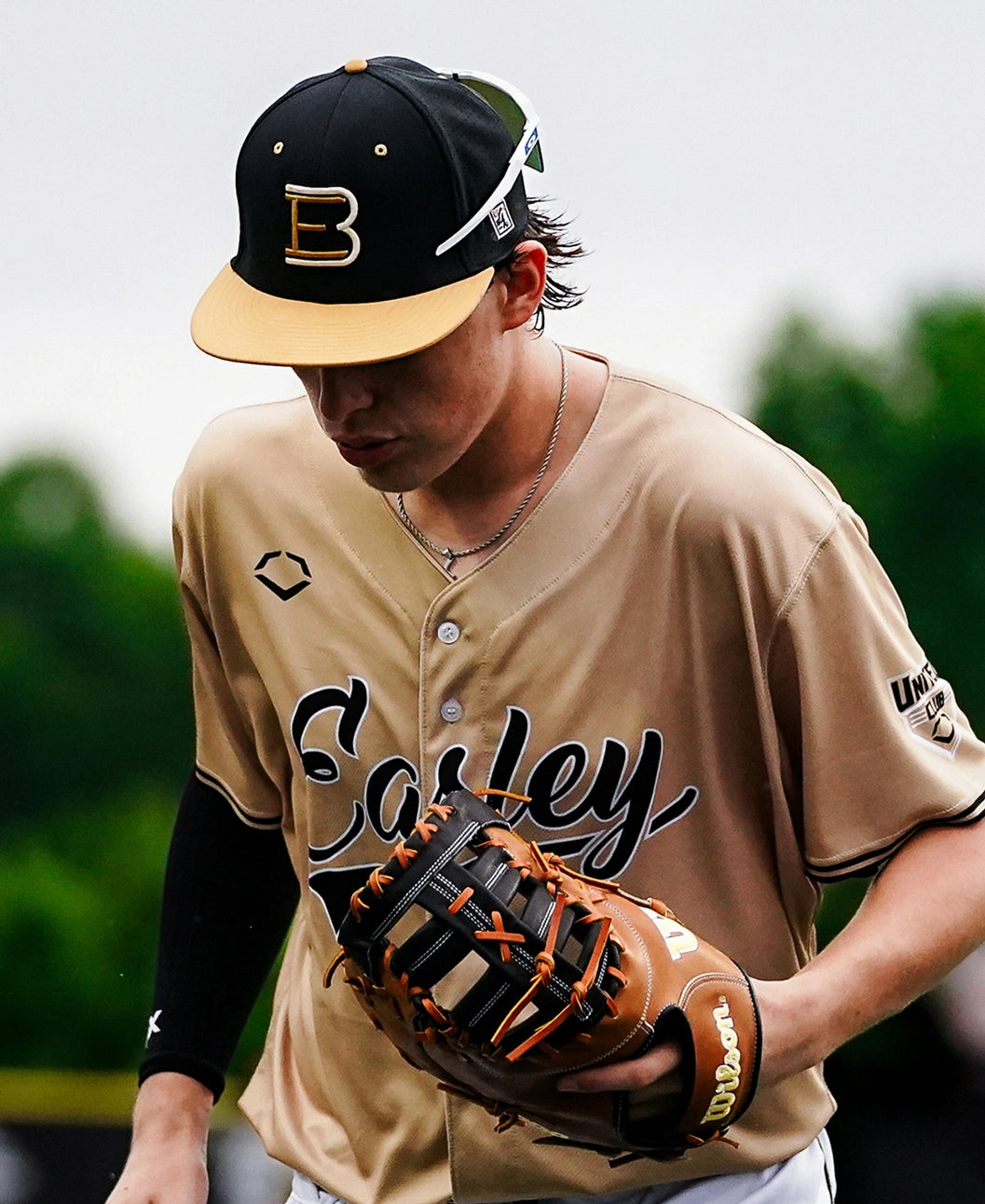 A baseball player in a tan Easley jersey and black cap holds a Pro Stock leather first base mitt while looking down. The background is blurred greenery, suggesting an outdoor field.