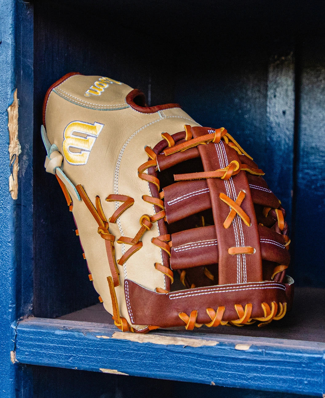 A tan and brown 13 inch glove crafted from Pro Stock leather with orange laces rests on a blue wooden shelf, ready for use in a game.