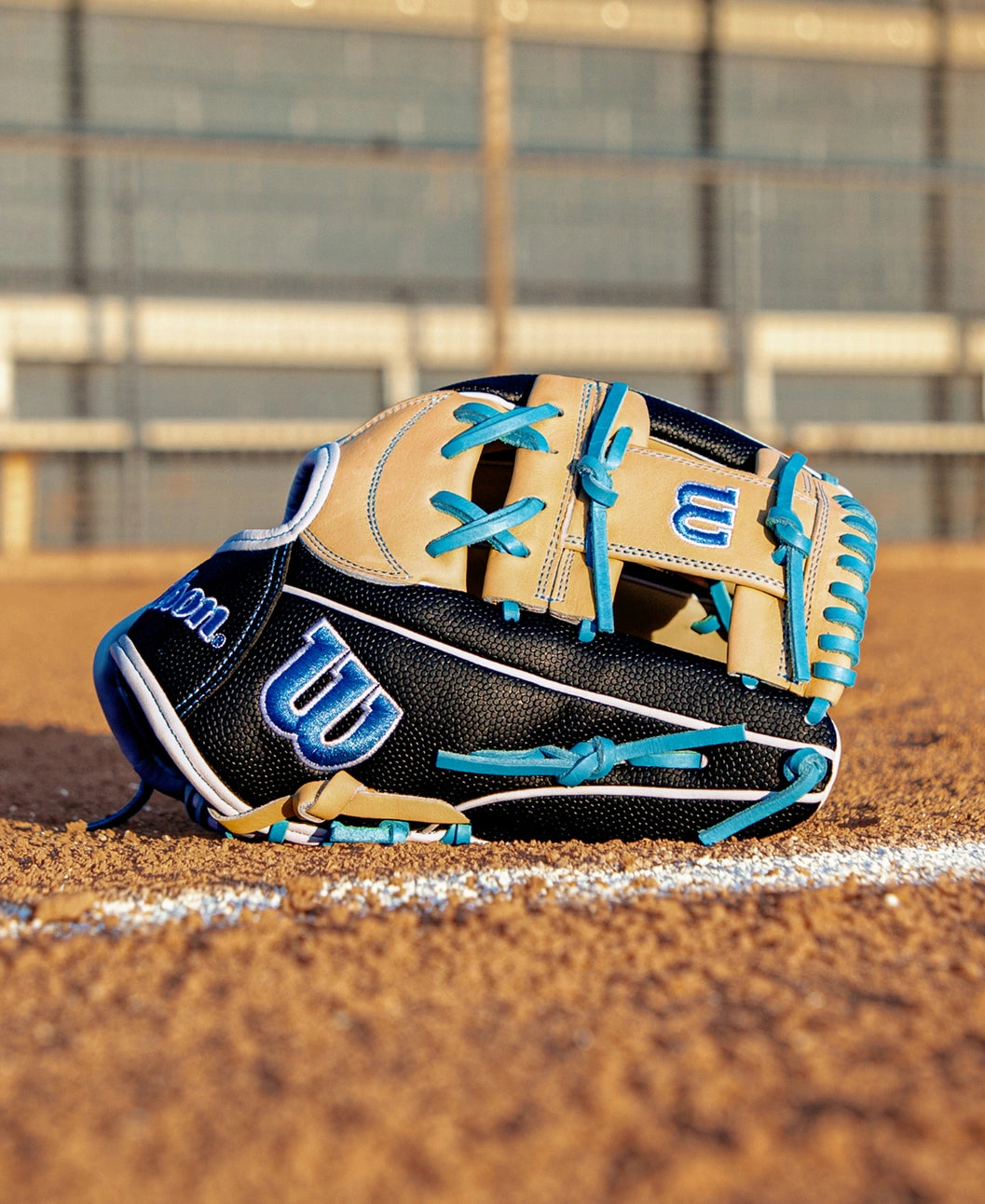 A Wilson A2000 FP1175SS 11.75″ Fastpitch Glove (WBG06601RT1175) in black and tan with blue laces rests on a dirt field near a white foul line, with a fence blurred in the background.