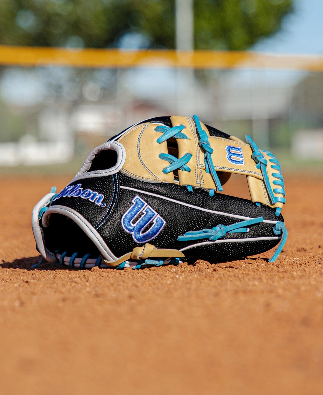 A Wilson A2000 FP1175SS 11.75″ Fastpitch Glove (WBG06601RT1175) by Wilson, featuring a black and tan design with blue laces, rests on a dirt baseball field with a blurred fence and trees in the background.