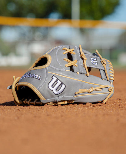 A Wilson A2000 H12 12″ Fastpitch Glove (WBG06801RT12) in gray with tan laces sits on a dirt infield, while a blurred fence, yellow barrier, and green trees fill the background.