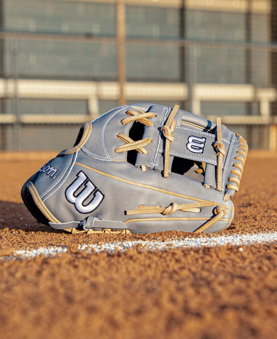 A Wilson A2000 H12 12″ Fastpitch Glove: WBG06801RT12 by Wilson in gray lies on the dirt near a white chalk line on a baseball field, with out-of-focus fencing and stands visible in the background.