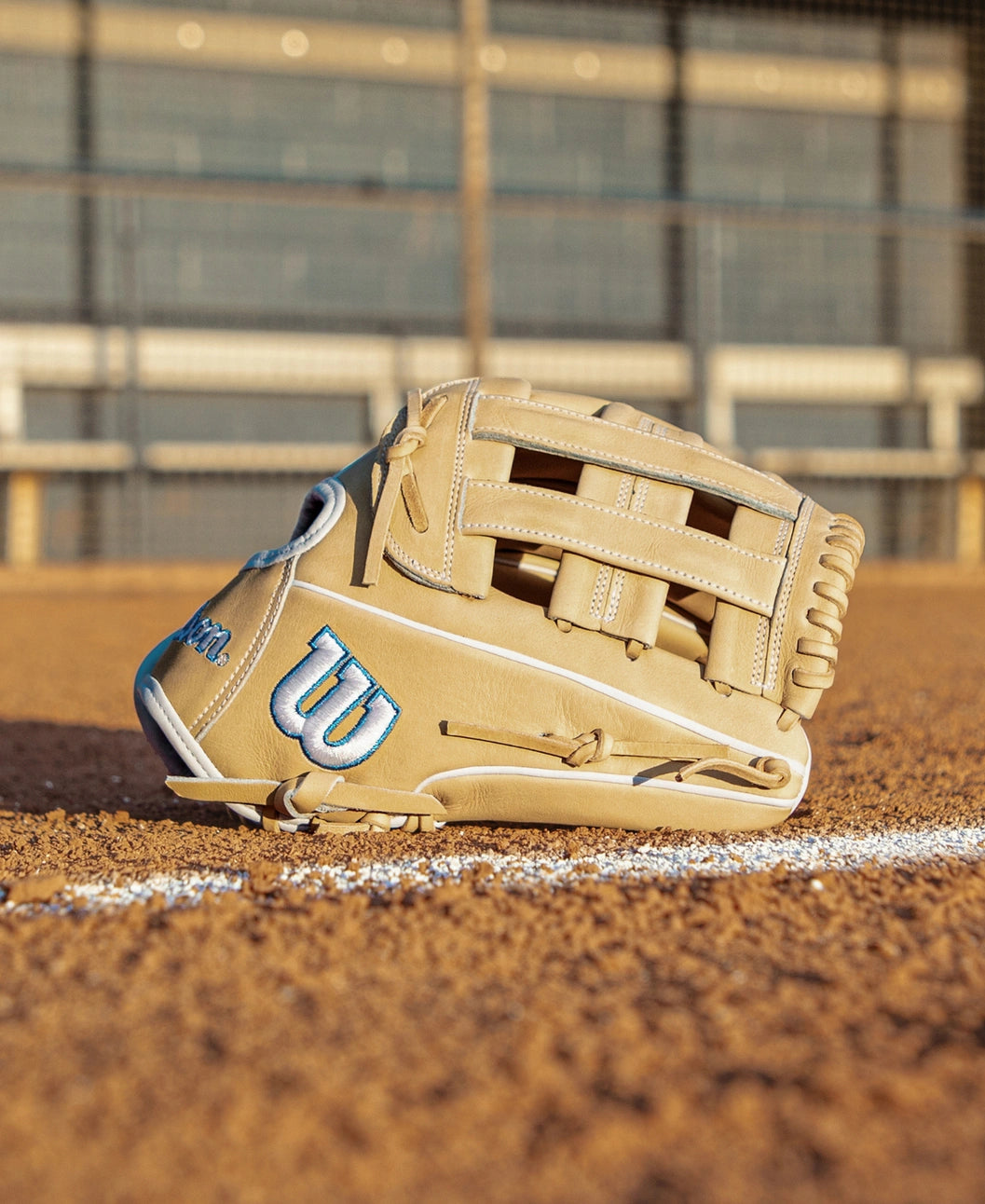A Wilson A2000 FP1712 12″ Fastpitch Glove (WBG06901RT12) by Wilson, in tan, rests on a dirt field near a white chalk line, with empty bleachers and a fence visible in the blurred background.