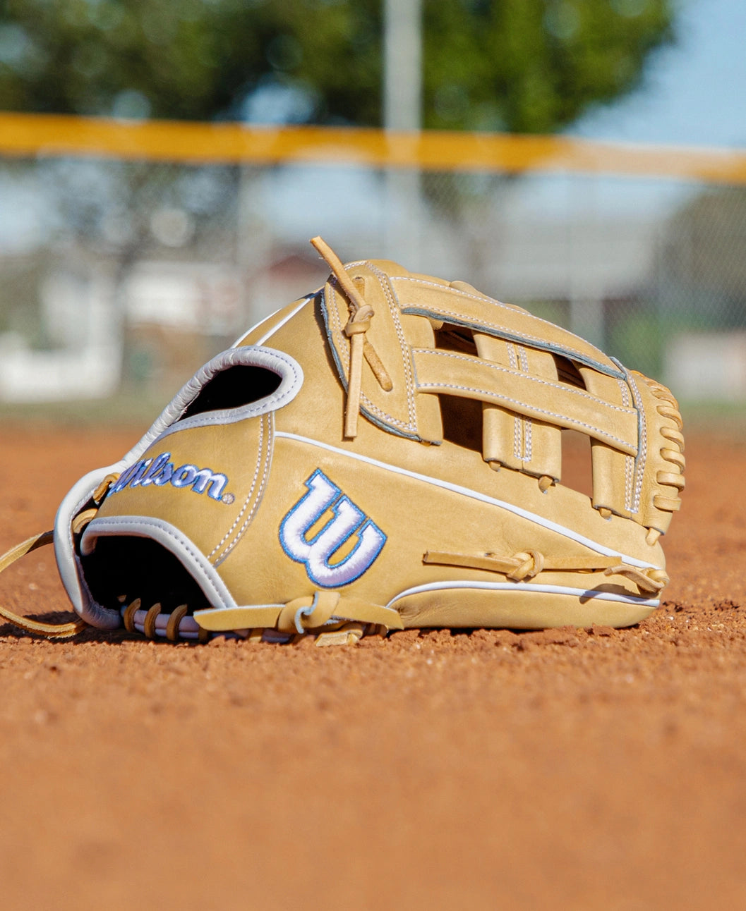 A Wilson A2000 FP1712 12″ Fastpitch Glove (WBG06901RT12) in tan, featuring blue and white stitching, rests on a dirt baseball field with a blurred fence and trees in the background.