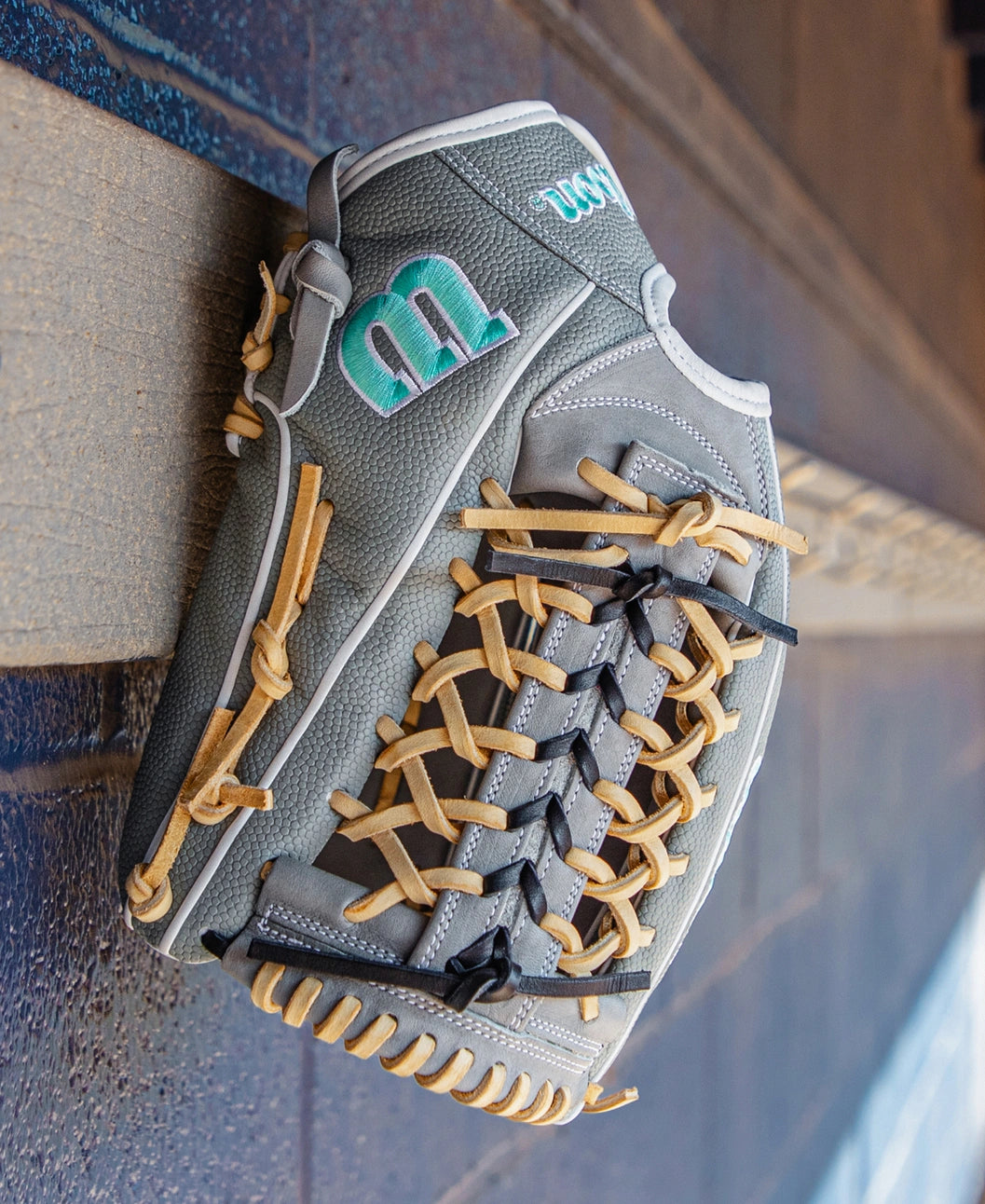 A Wilson A2000 T125SS 12.5″ Fastpitch Glove (WBG07001RT125) with gray leather, tan laces, and a green M logo hangs on a wooden ledge against a dark brick wall.