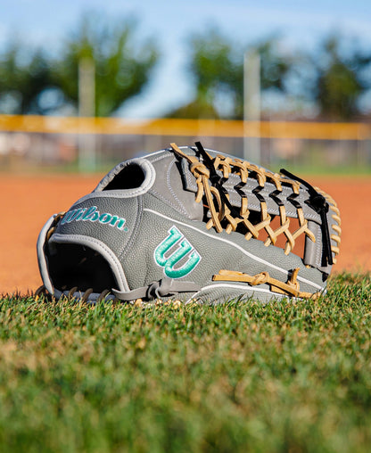 A Wilson A2000 T125SS 12.5″ Fastpitch Glove (WBG07001RT125) by Wilson, featuring a gray and tan design, rests on green grass near a dirt infield, with a blurred baseball field and trees in the background beneath a blue sky.