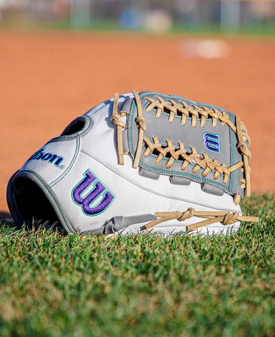 A Wilson A2000 FPHGSS 12″ Fastpitch Glove (WBG07101RT12) by Wilson, featuring a white and gray design with purple and teal accents, rests on green grass near a dirt baseball field, highlighting its Flex System for the ideal softball infield performance.