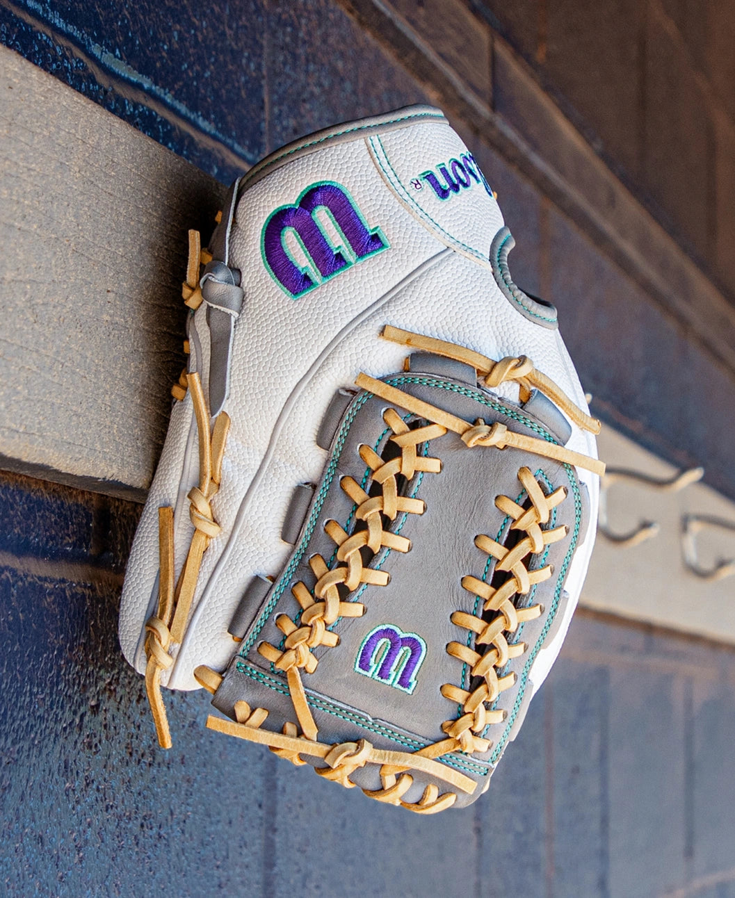 A Wilson A2000 FPHGSS 12″ Fastpitch Glove (WBG07101RT12) by Wilson, in white and gray with tan laces and accented with purple and teal M logos, hangs on a wooden wall with decorative trim. Designed for softball infielders, this glove incorporates the innovative Flex System to deliver enhanced comfort and performance.