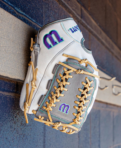 A Wilson A2000 FPHGSS 12″ Fastpitch Glove (WBG07101RT12) by Wilson, in white and gray with tan laces and accented with purple and teal M logos, hangs on a wooden wall with decorative trim. Designed for softball infielders, this glove incorporates the innovative Flex System to deliver enhanced comfort and performance.