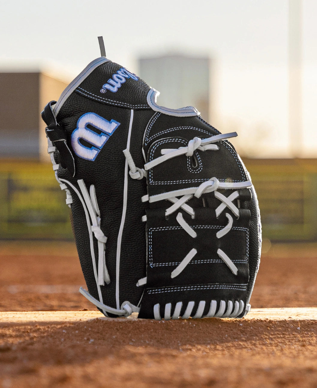 A Wilson A2000 FPB25SS 12.25″ Fastpitch Glove (WBG07201RT1225) stands upright on the dirt of a baseball field, its black leather accented with white laces, with blurred buildings and a fence in the background.
