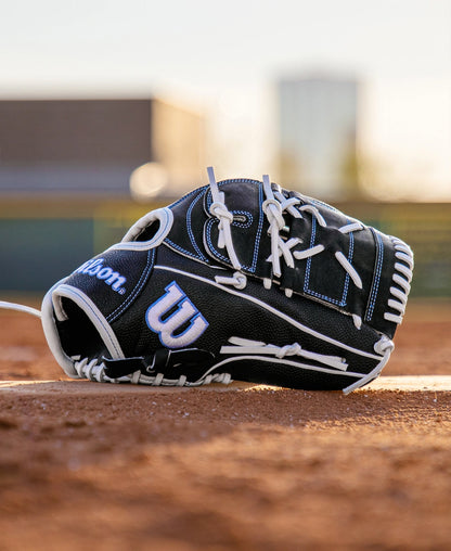A Wilson A2000 FPB25SS 12.25″ fastpitch glove (WBG07201RT1225) by Wilson, featuring a sleek black design with white laces, rests on the dirt of a baseball field, while the stadium and nearby buildings are softly blurred in the background.