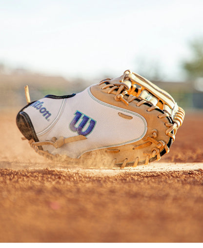 A Wilson A2000 FPCM23SS 34″ Fastpitch Catcher’s Mitt, featuring a white and tan design with a Dual Post Web, lies on a dusty field, kicking up dirt against a backdrop of blurred outdoor scenery.