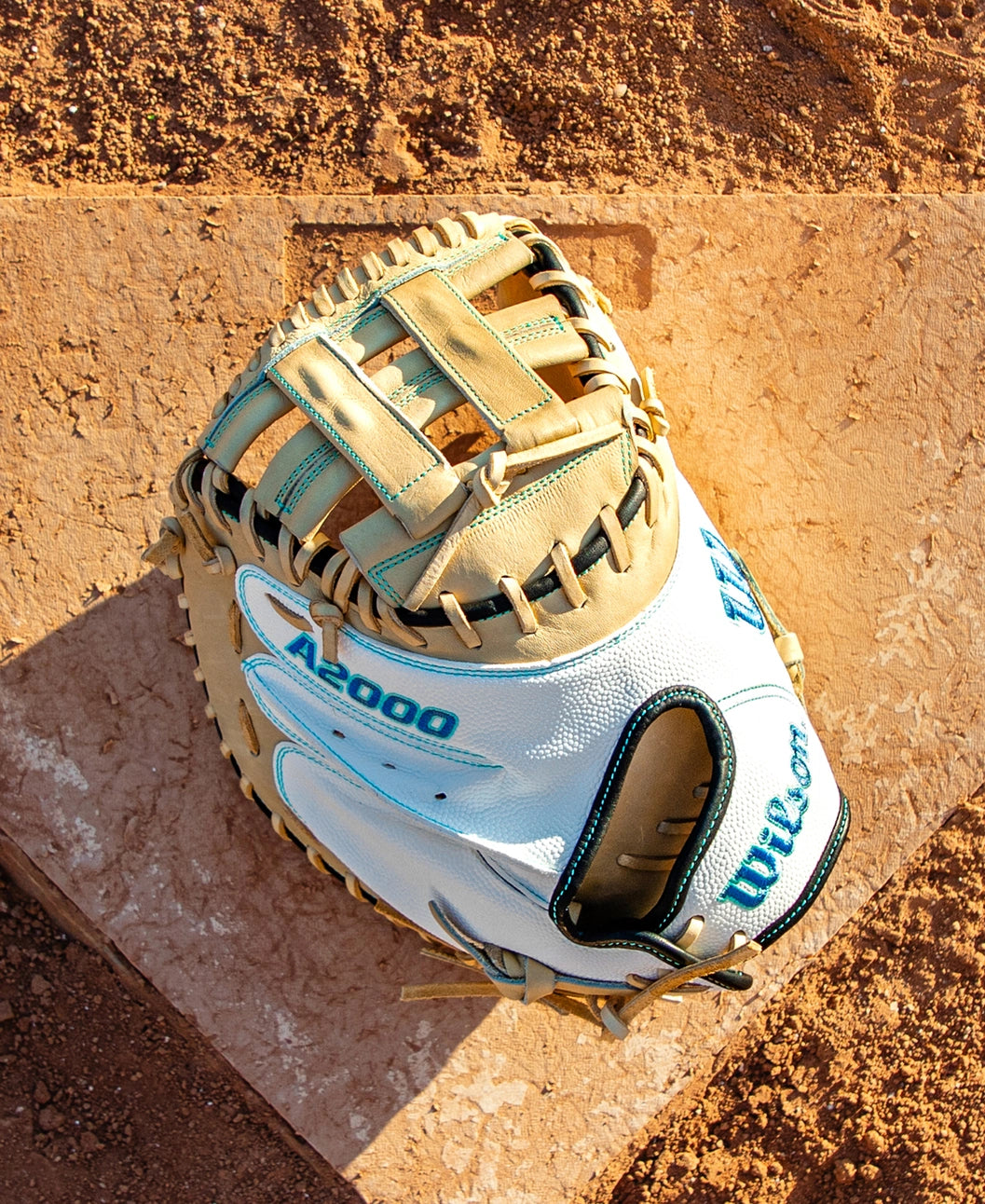 A Wilson A2000 FPCM23SS 34″ Fastpitch Catcher’s Mitt (WBG07301RT34) by Wilson, featuring a white and tan design, rests on a dusty home plate surrounded by brown dirt on a sunny day.