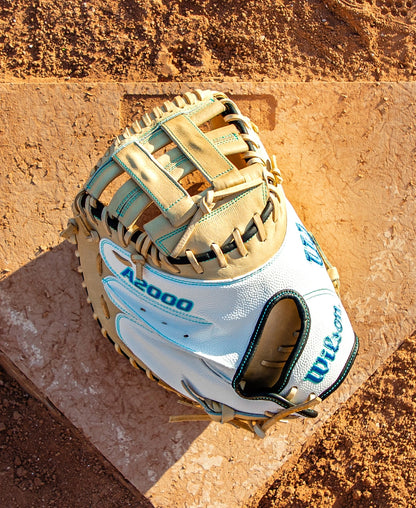 A Wilson A2000 FPCM23SS 34″ Fastpitch Catcher’s Mitt (WBG07301RT34) by Wilson, featuring a white and tan design, rests on a dusty home plate surrounded by brown dirt on a sunny day.