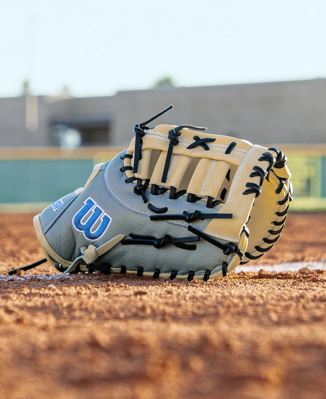 A Wilson A2000 FP1620SS 12.5″ Fastpitch First Base Mitt (WBG07401RT125) made from Pro Stock leather rests on the infield dirt of a baseball field, with a blurred building and green fence in the background.
