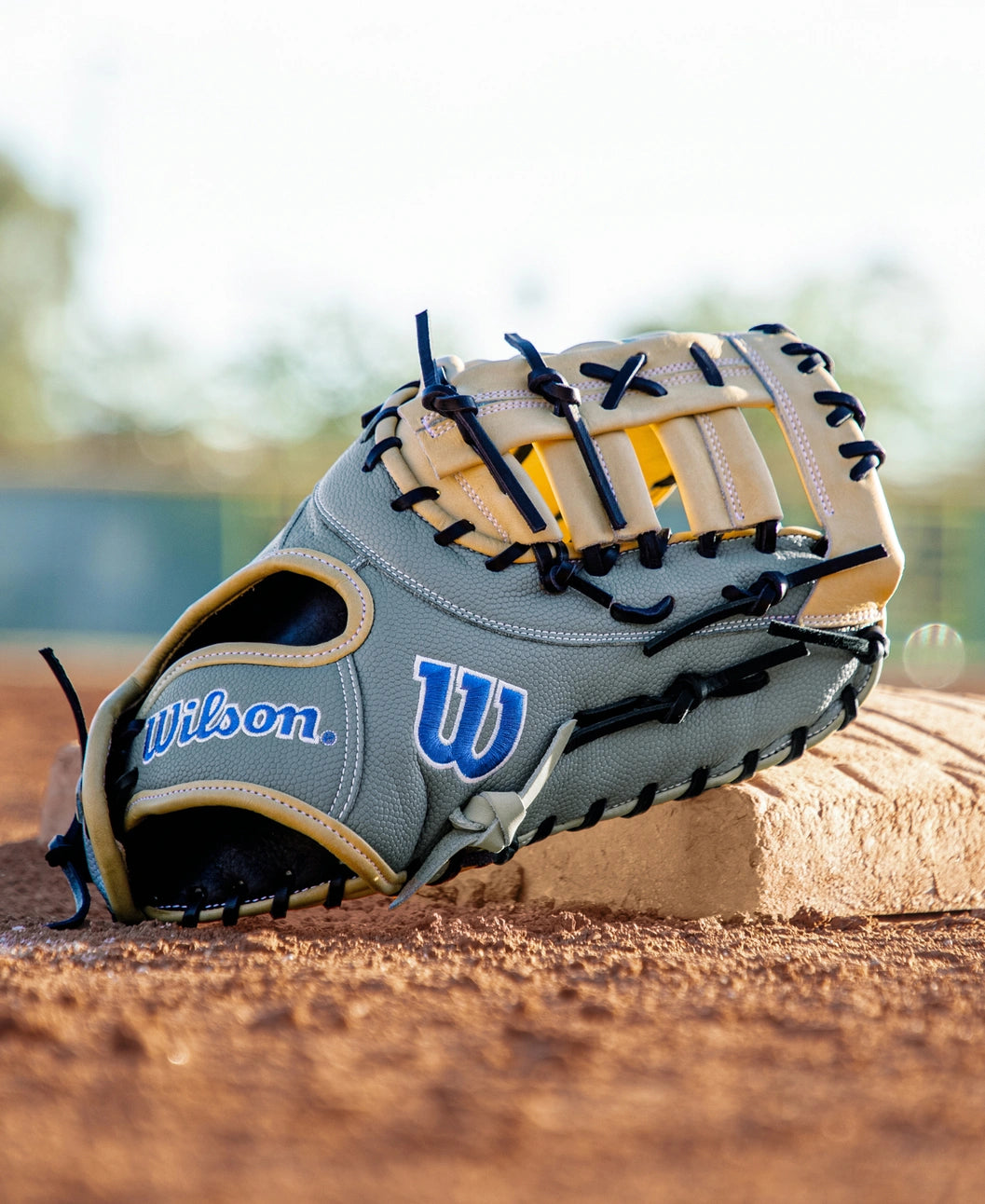 A Wilson A2000 FP1620SS 12.5″ Fastpitch First Base Mitt (WBG07401RT125) rests on a pitcher’s mound on a baseball field, its Pro Stock leather and tan and black laces standing out against the backdrop of blurred green fencing and trees.