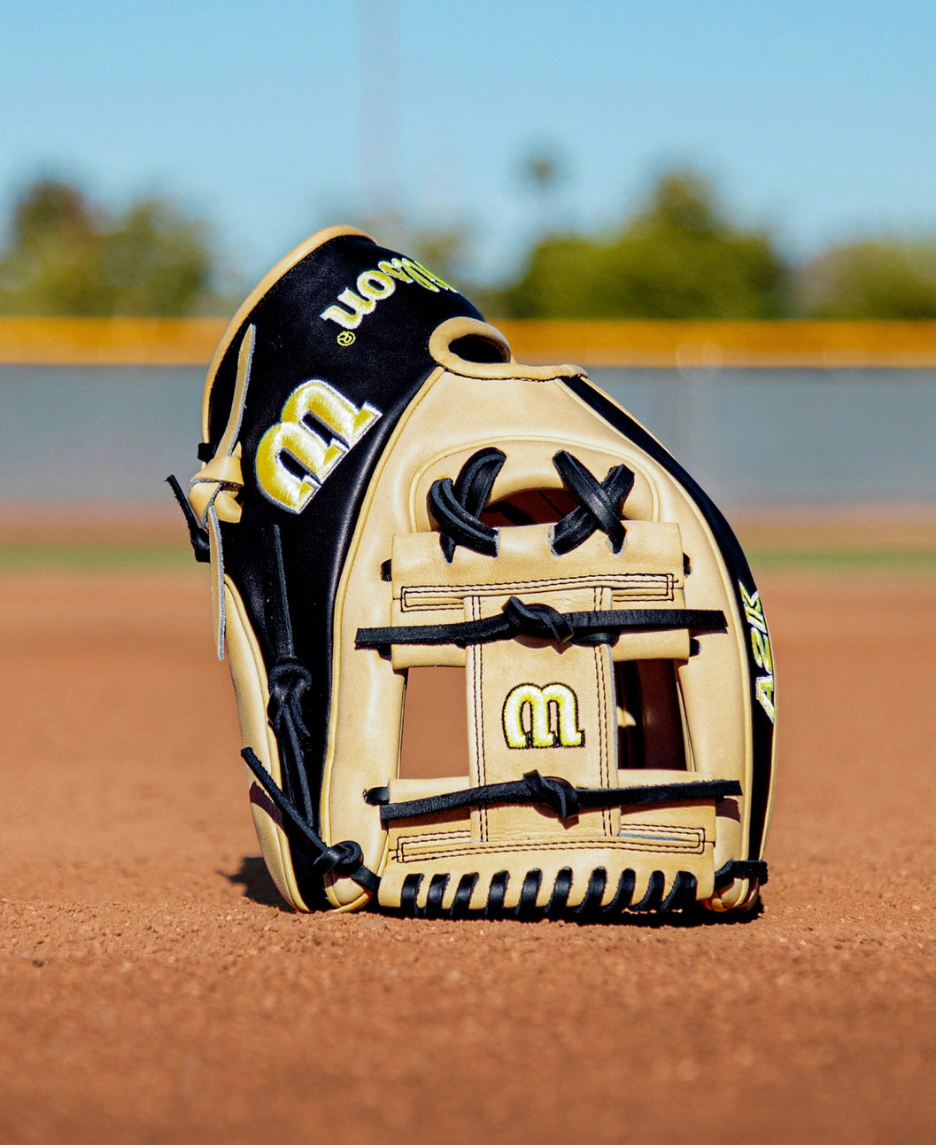 A Wilson A2K 1786 11.5″ Baseball Glove (WBG07601RT115) in tan and black is displayed upright on a dirt infield, with a blurred outfield fence and trees in the background under clear daylight.