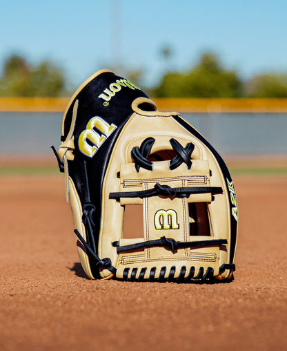 A Wilson A2K 1786 11.5″ Baseball Glove (WBG07601RT115) in tan and black is displayed upright on a dirt infield, with a blurred outfield fence and trees in the background under clear daylight.