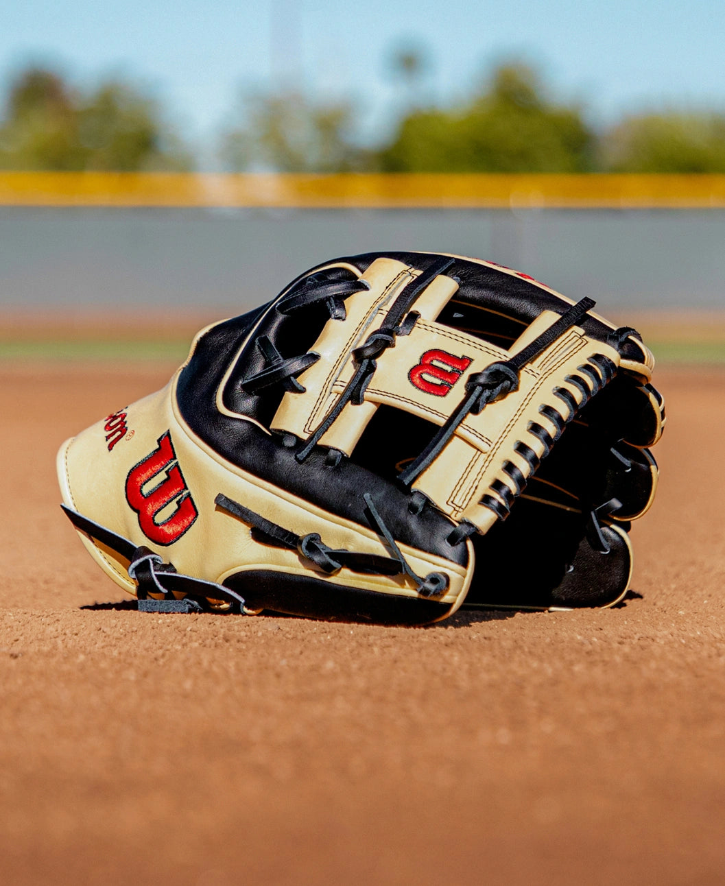A Wilson A2K 1724 11.75″ Baseball Glove (WBG07701RT1175), in tan and black with red Wilson logos, lies on the infield dirt of a baseball field, highlighting its Double Palm Construction; blurred grass and a yellow outfield fence can be seen in the background.