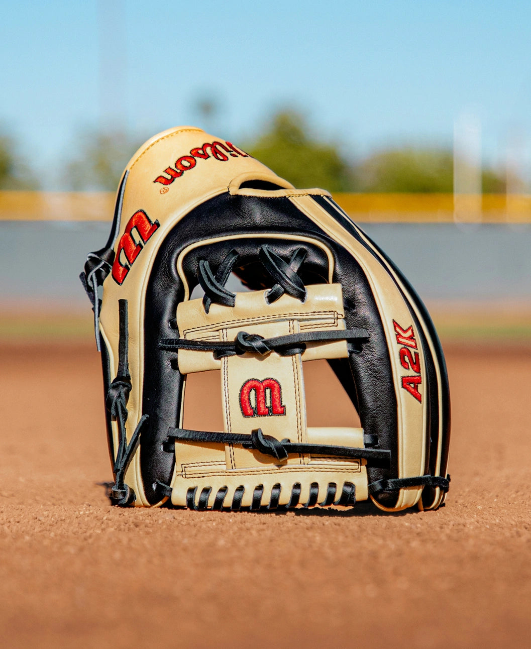 A Wilson A2K 1724 11.75″ Baseball Glove (WBG07701RT1175) by Wilson stands upright on a dirt baseball field, highlighting its Double Palm Construction, with a blurred fence and trees in the background beneath a clear blue sky.
