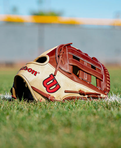 A Wilson A2K DW5 12″ Baseball Glove (WBG07801RT12) by Wilson, featuring brown and tan Pro Stock Select leather, lies on green grass with a blurred fence and blue sky behind it.
