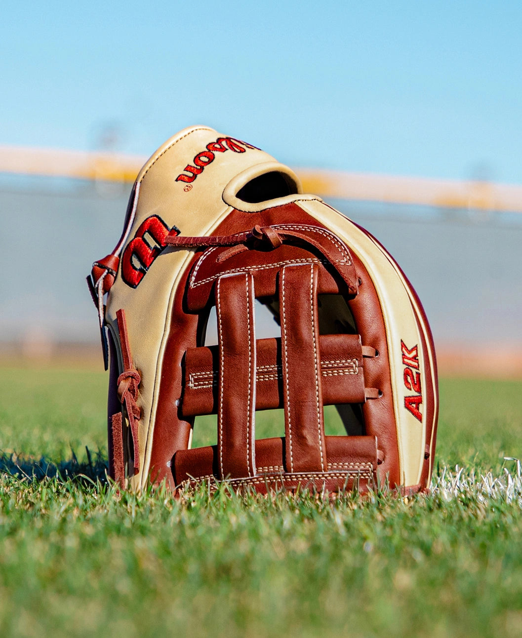 A Wilson A2K DW5 12″ Baseball Glove (WBG07801RT12), featuring tan and brown Pro Stock Select leather, stands upright on green grass with a blurred stadium backdrop beneath a clear blue sky.
