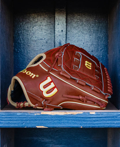A Wilson A2K C12 12″ Baseball Glove (WBG07901RT12) by Wilson, featuring a rich brown color with yellow accents, rests on a wooden shelf, its deep pocket subtly highlighted against a dark blue background.