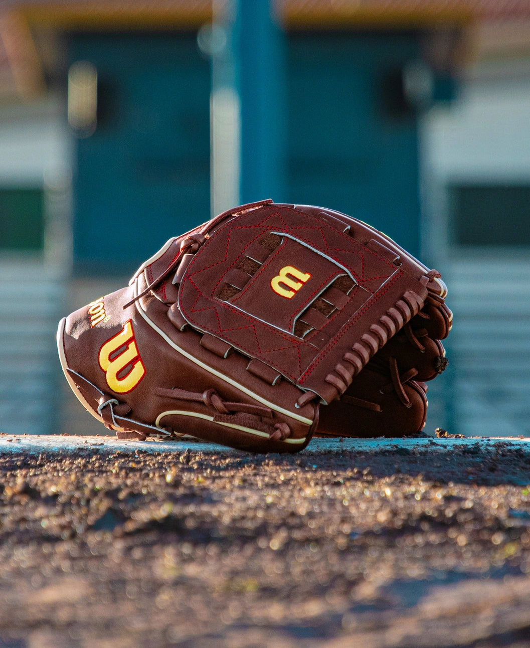 A Wilson A2K C12 12″ Baseball Glove (WBG07901RT12) made of brown leather with yellow stitching and featuring the Wilson “W” logo rests on the dirt of a baseball field, its deep pocket ready for action, while blurred bleachers fill the background.