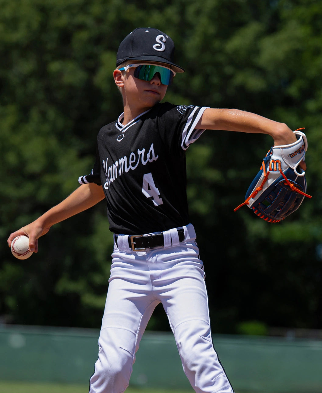 A young baseball player wearing a team uniform and mirrored sunglasses winds up to throw a ball while wearing a Wilson A500 10.5″ Baseball Glove: WBG08401RT105 all-leather youth glove.
