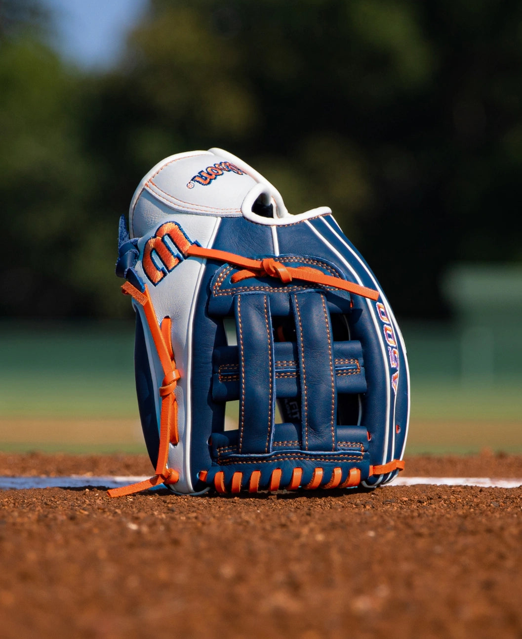 A blue and white Wilson A500 10.5″ Baseball Glove: WBG08401RT105 with orange laces standing upright on a dirt field.