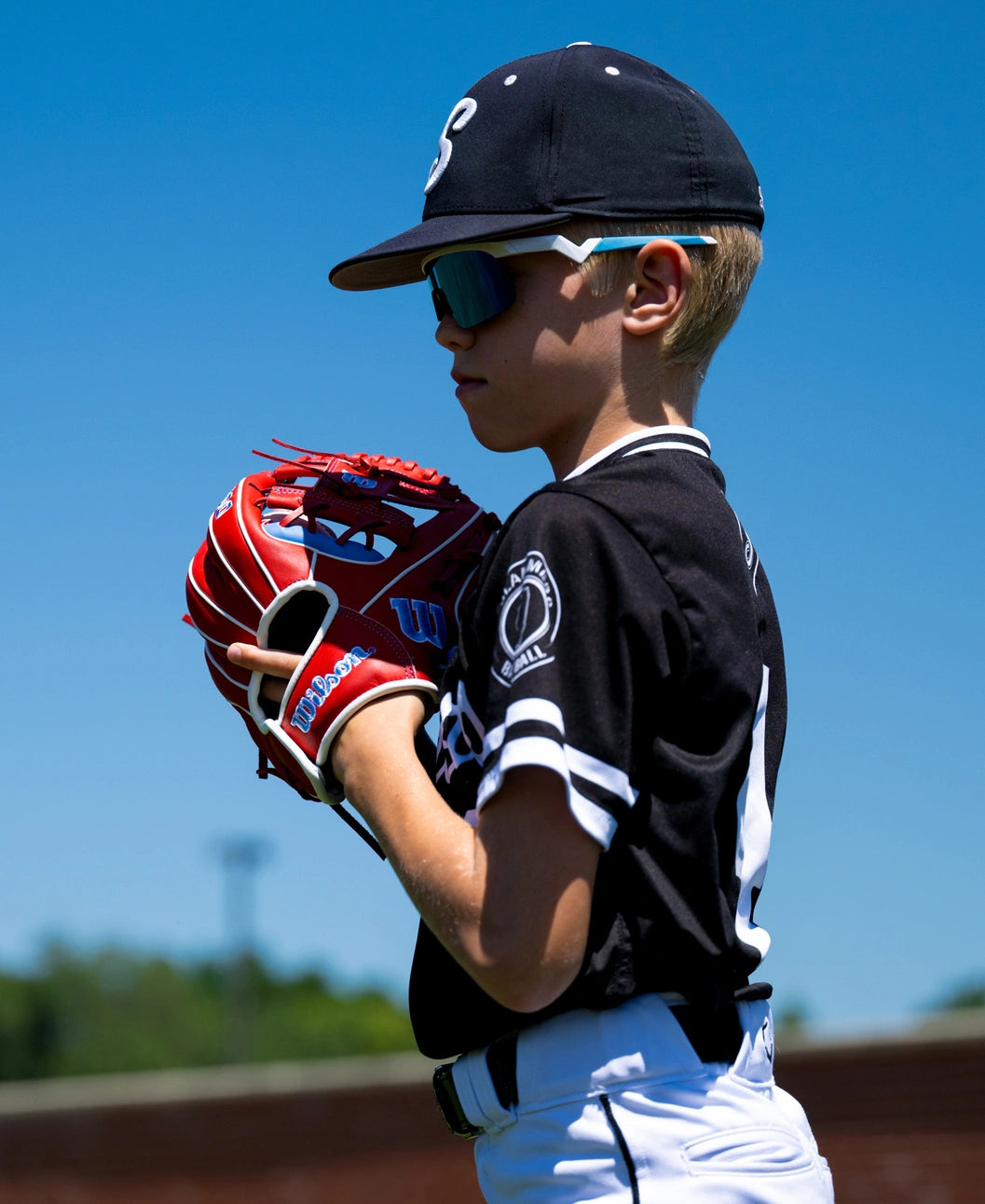 A young baseball player wearing a cap and sunglasses holds a red and white Wilson A500 11″ Baseball Glove: WBG08501RT11.