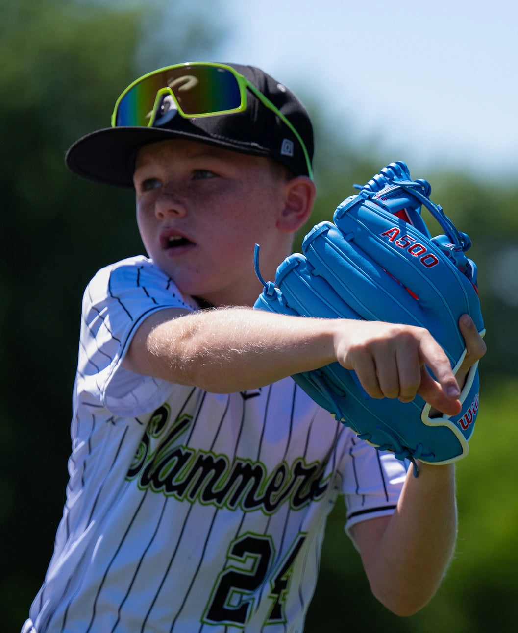 A youth baseball player in a pinstriped uniform wears a bright blue Wilson A500 11.5″ Baseball Glove: WBG08601RT115 while pointing.