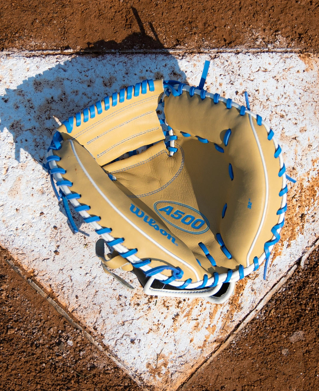 A tan Wilson A500 32″ Baseball Catcher’s Mitt: WBG08901RT32 with bright blue lacing, resting on a white home plate.