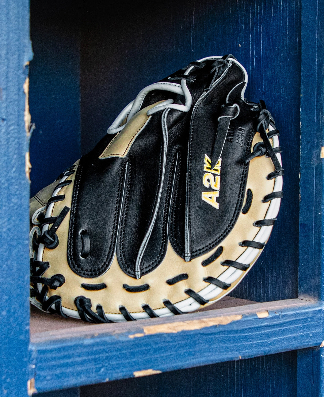 A Wilson A2K 1734 34″ black and tan baseball catcher’s mitt with Double Palm Construction rests on a blue wooden shelf with chipped paint.