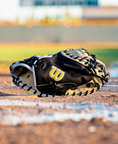 A Wilson A2K 1734 34″ Baseball Catcher’s Mitt (WBG10701RT34) in black and tan rests on the ground near home plate on a baseball field, with grass and a building softly blurred in the background.
