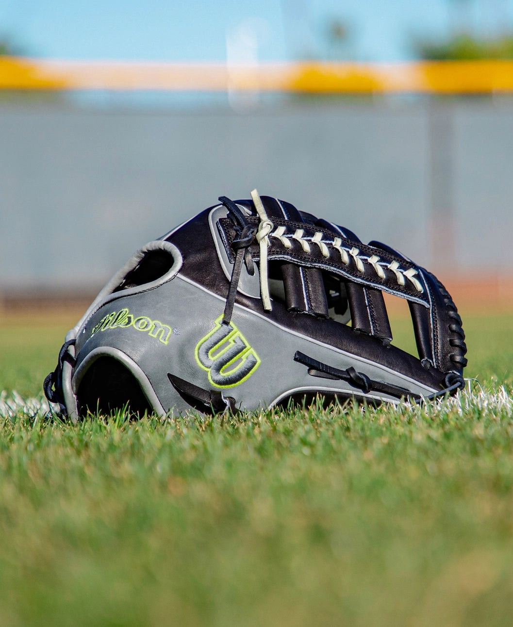 A black and gray Wilson A2K 1810 12.75″ Baseball Glove (WBG10802RT1275) rests on the grass near a white chalk line, with a blurred fence and yellow barrier in the background. Crafted by Wilson from Pro Stock Select leather, this glove offers exceptional durability and premium on-field performance.