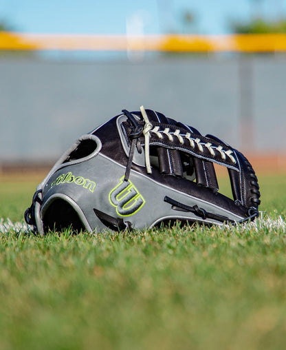 A black and gray Wilson A2K 1810 12.75″ Baseball Glove (WBG10802RT1275) rests on the grass near a white chalk line, with a blurred fence and yellow barrier in the background. Crafted by Wilson from Pro Stock Select leather, this glove offers exceptional durability and premium on-field performance.