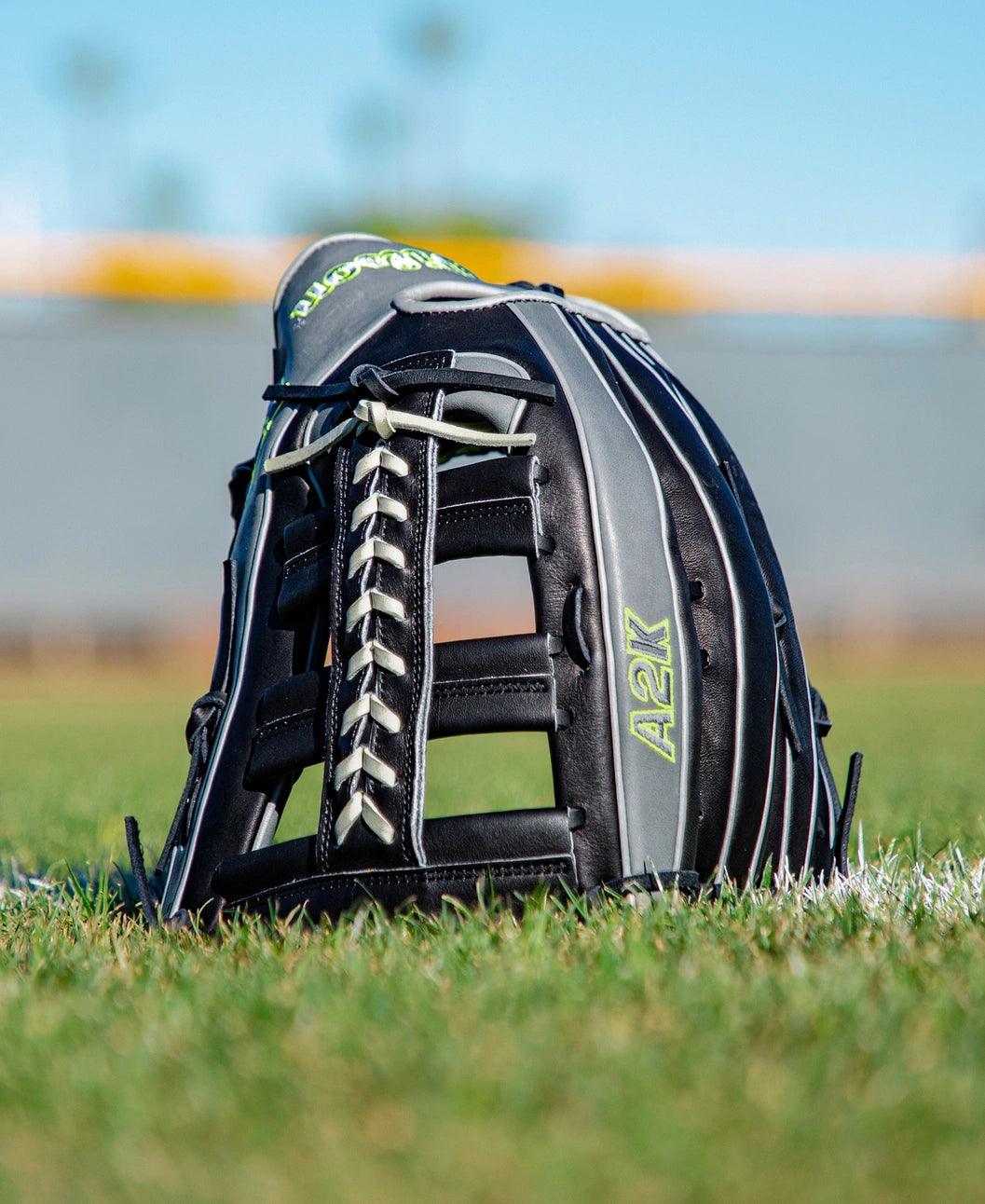 A Wilson A2K 1810 12.75″ Baseball Glove (WBG10802RT1275) from Wilson, featuring black Pro Stock Select leather and white laces with “A2K” written in yellow, sits upright on green grass with a blurred field and blue sky in the background.
