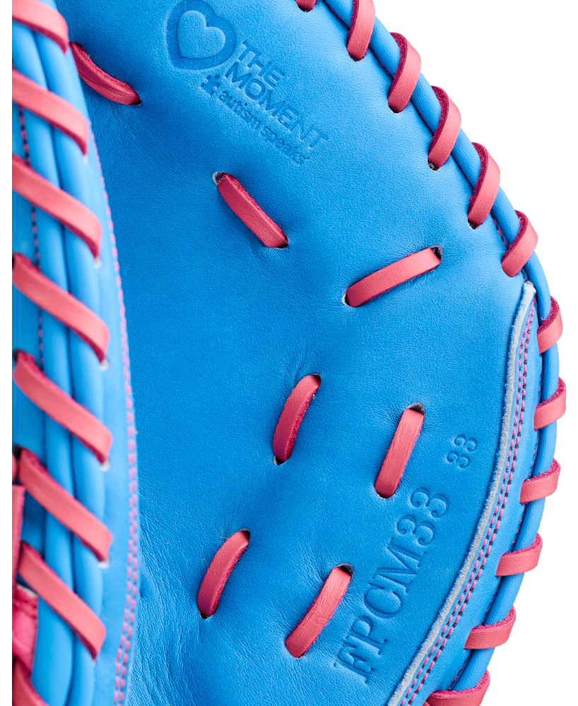 Close-up of a vibrant blue Wilson A1000 FPCM33 MOMENTUM 33″ Fastpitch Catcher’s Mitt, expertly crafted from Full Grain leather with pink laces and featuring embossed text reading “The Moment A Catch Signs” and “FPCM33 33” circumference.”.