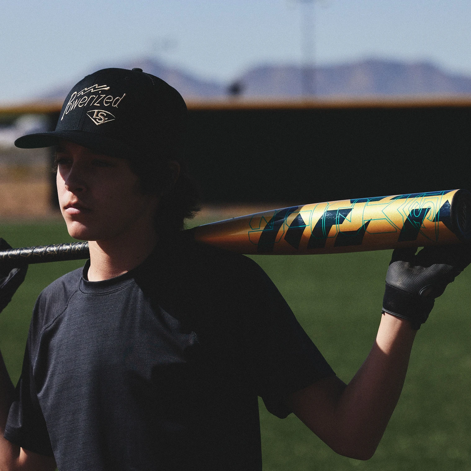 A young baseball player wearing a black shirt, black gloves, and a black cap rests a colorful bat on his shoulder while standing on a baseball field with mountains in the background.